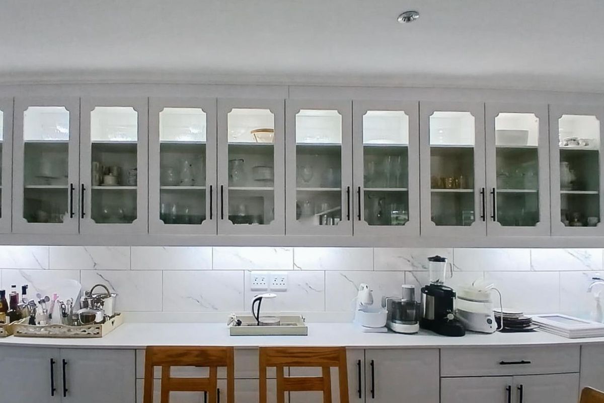 A well-lit kitchen featuring white cabinetry with glass-fronted upper cupboards, marble-look tiled backsplash, and integrated under-cabinet lighting.