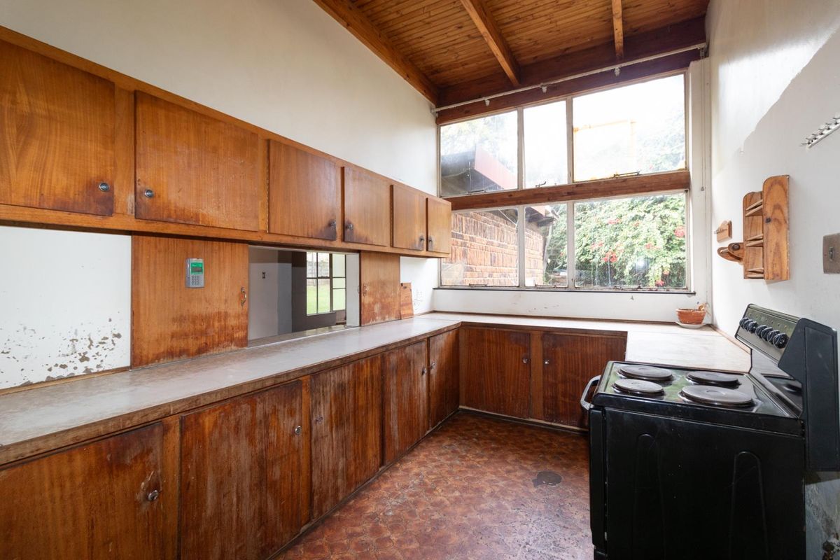 A dated kitchen featuring original timber cabinetry, a freestanding stove, and high windows with exposed timber ceiling beams, offering significant potential for renovation.