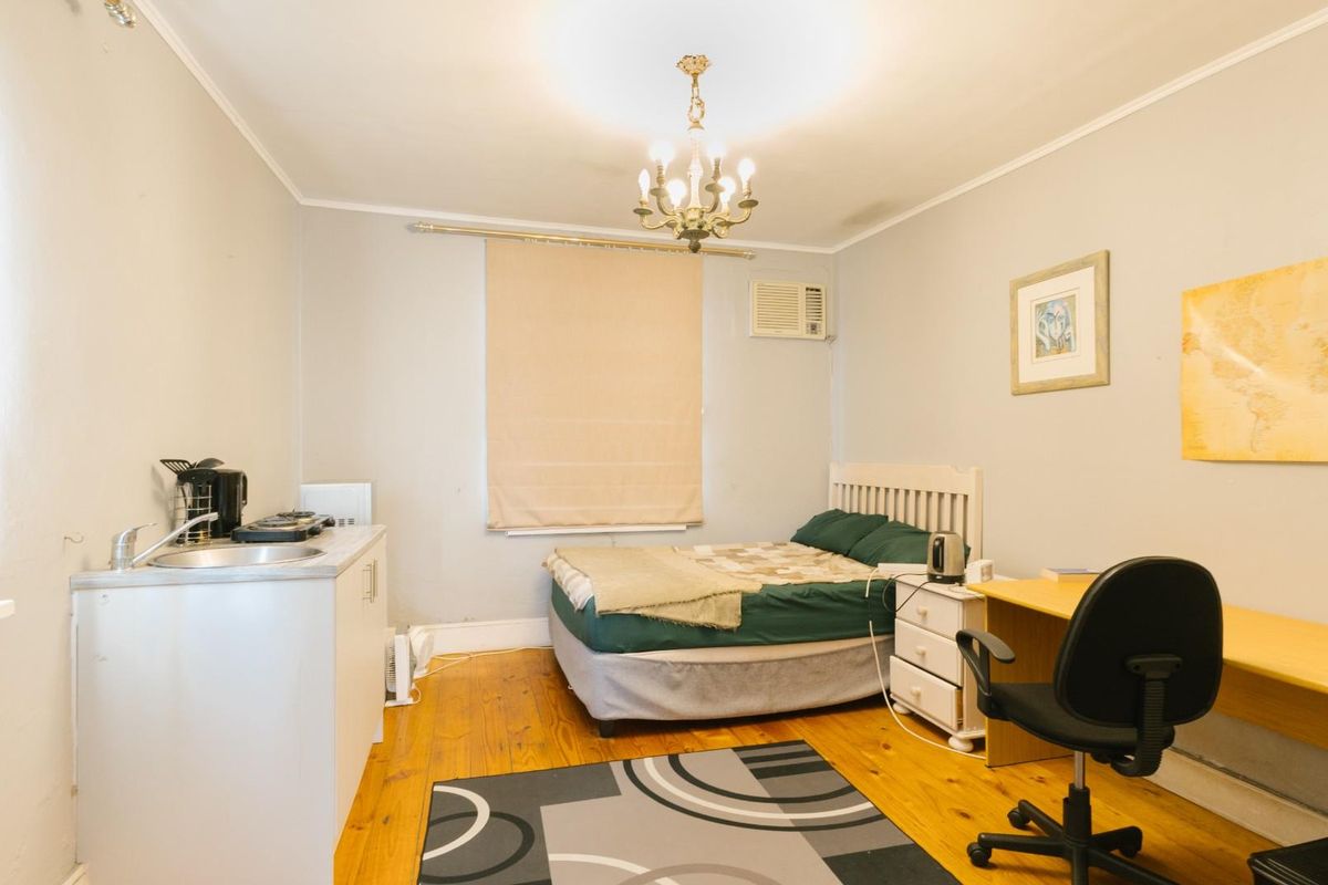 A studio-style bedroom featuring timber flooring, a compact kitchenette unit with sink, and a wall-mounted air conditioning unit.