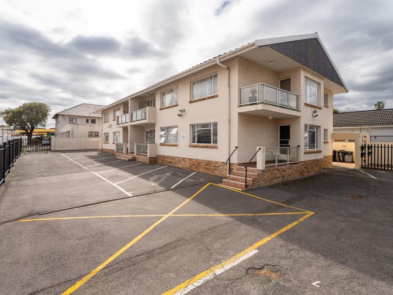 Exterior view of a multi-story apartment building featuring a paved parking area, brick base detailing, and private balconies.