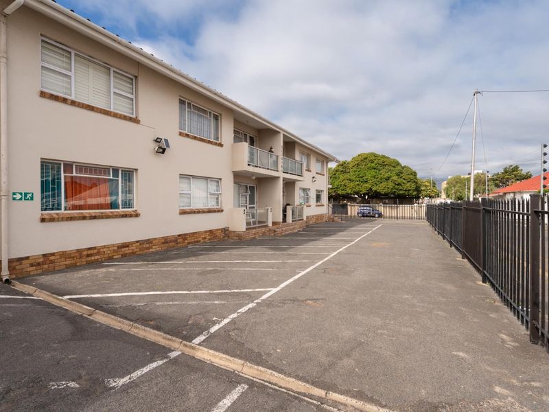 Exterior view of an apartment complex featuring a paved parking area, metal perimeter fencing, and multiple balconies with sliding windows.