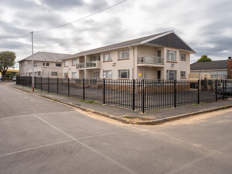 Exterior view of a multi-story apartment complex featuring a paved parking area, secure metal perimeter fencing, and private balconies.