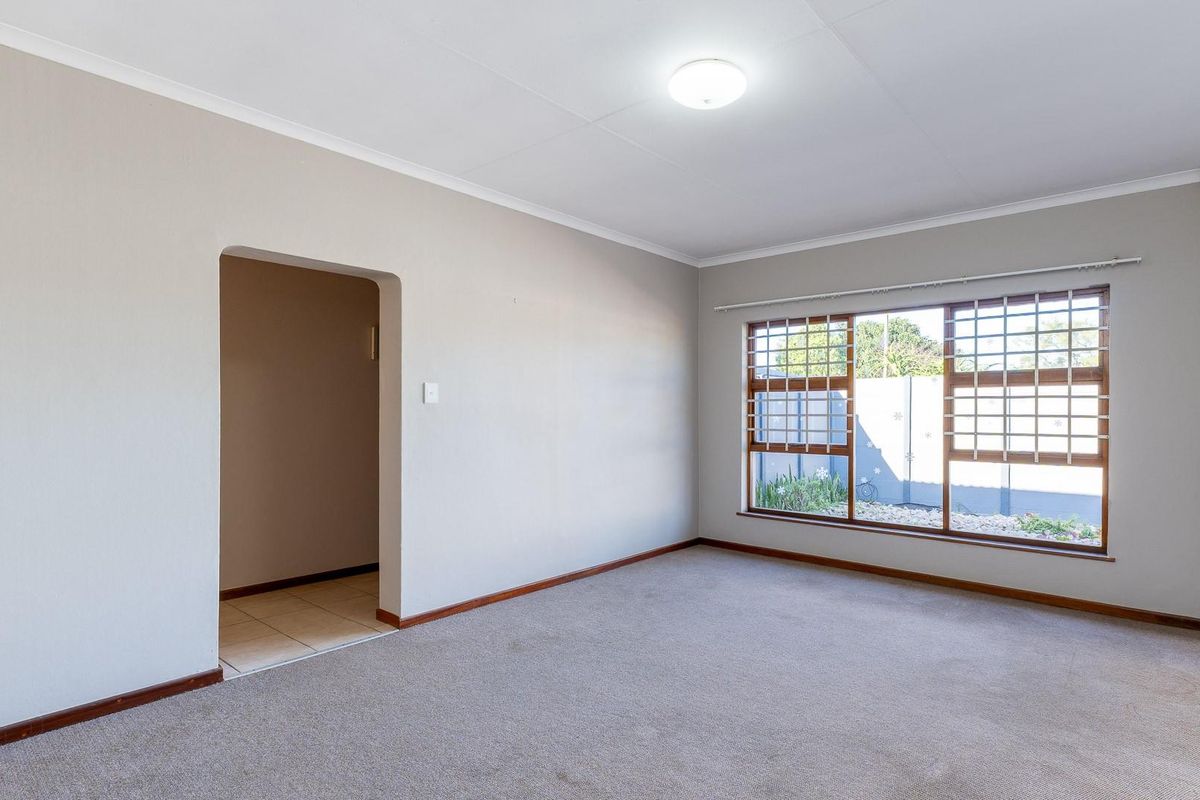 A spacious, empty living room featuring neutral walls, carpeted flooring, and large windows with security bars, offering a blank canvas for personalization.