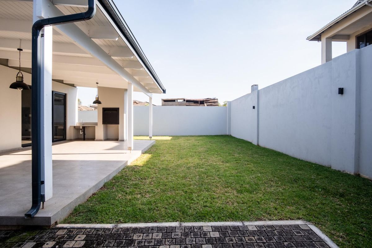 A spacious rear garden featuring a covered patio with a built-in braai area, paved sections, and a well-maintained lawn enclosed by perimeter walls.