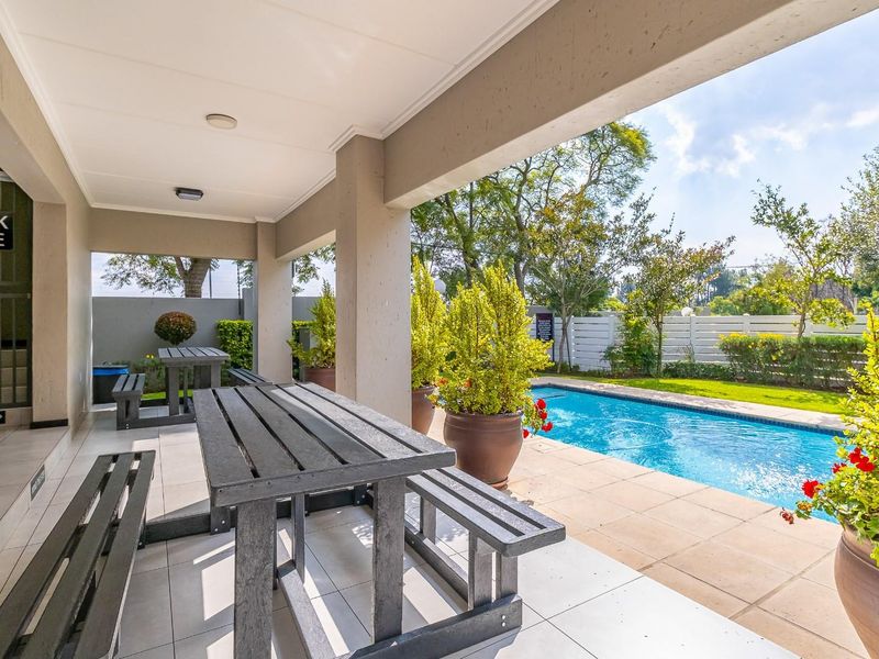 A covered patio area overlooking a swimming pool and garden, featuring tiled flooring and structural pillars, ideal for outdoor relaxation and entertainment.