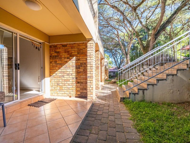 A covered patio area with tiled flooring, leading to a paved walkway and external staircase with metal railings, set within a landscaped garden.