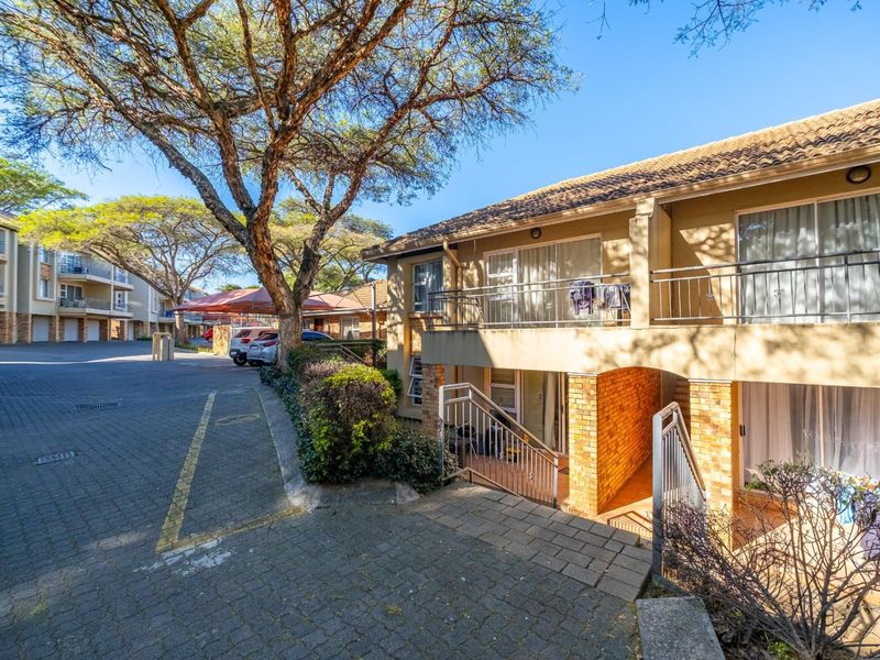 Exterior view of a townhouse complex featuring paved driveway, covered parking, and a multi-level building with balconies and brick accents.