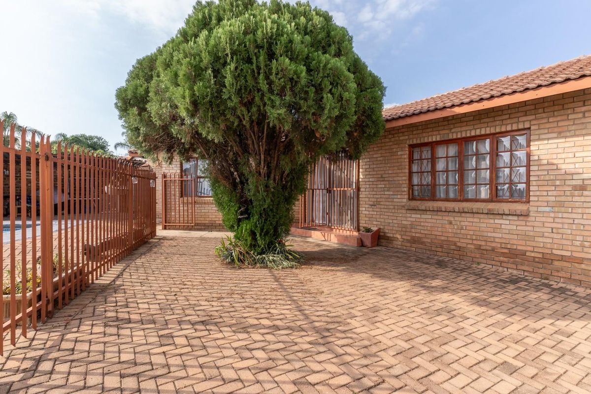 A paved rear courtyard area featuring face-brick exterior walls, security gates, and a mature tree, with a glimpse of the adjacent pool area.
