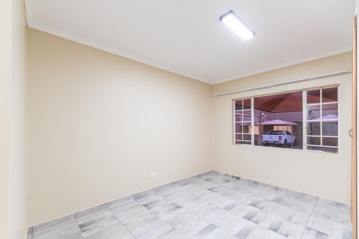 A bright, empty bedroom featuring neutral wall paint, tiled flooring, and a large window with security bars overlooking the exterior.