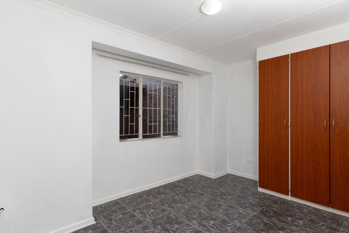 A clean, empty bedroom featuring tiled flooring, built-in wooden cupboards, and a window fitted with security bars.