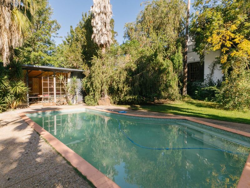 A private backyard featuring a swimming pool, surrounding paved area, and mature garden landscaping with a small detached structure.