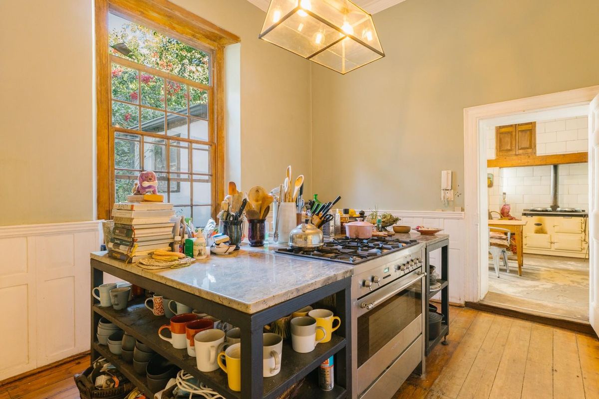 A rustic kitchen featuring a central island with a built-in gas hob and oven, timber flooring, and a view into an adjacent scullery area.