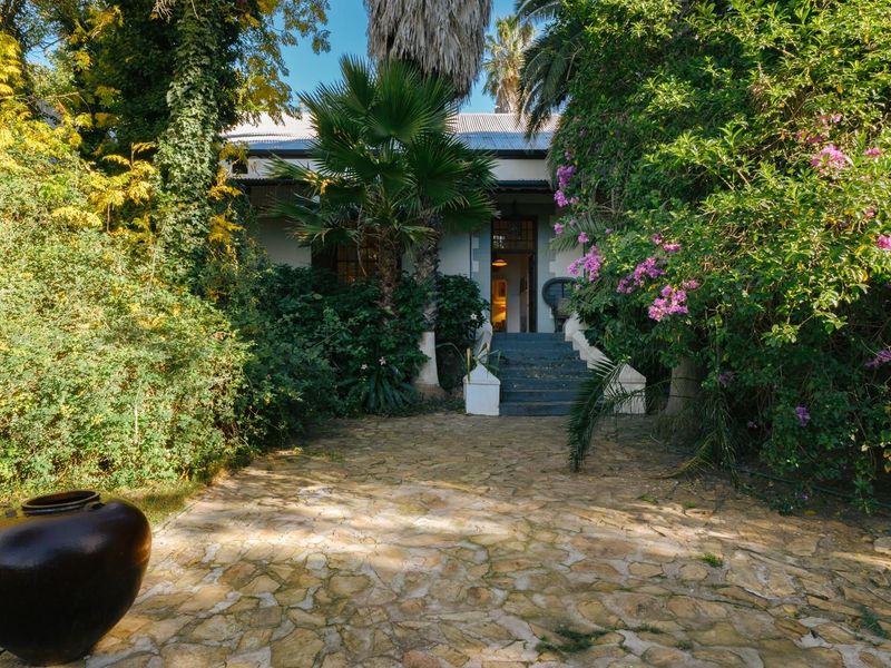 A charming front entrance featuring a stone-paved pathway leading to a raised porch, surrounded by lush, mature garden landscaping.