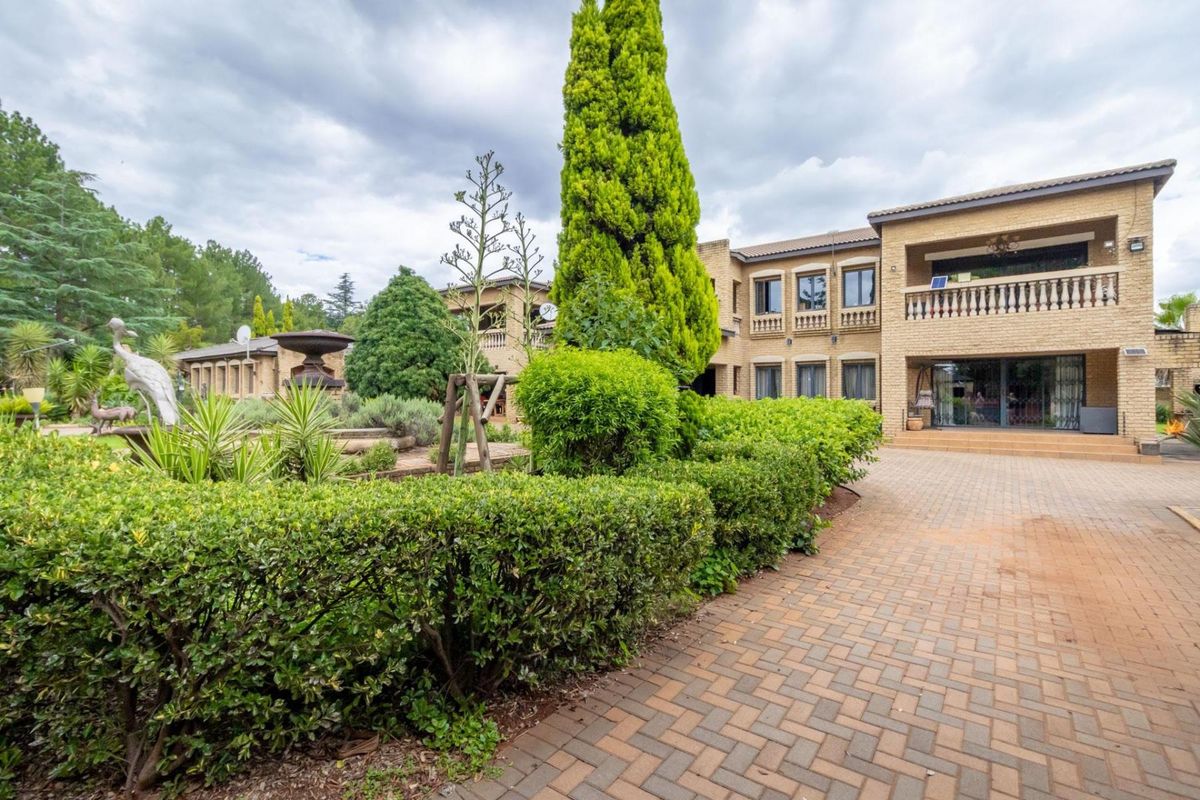 A spacious exterior rear view featuring a paved driveway, landscaped garden with mature hedges, and a multi-story brick residence with a balcony.