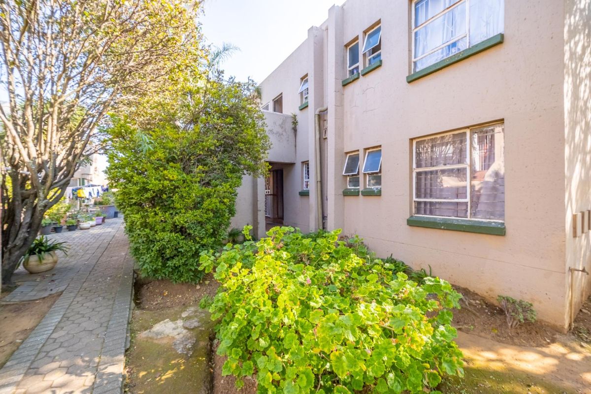 Rear exterior view of the apartment building featuring a paved walkway, landscaped garden area, and multiple casement windows along the facade.