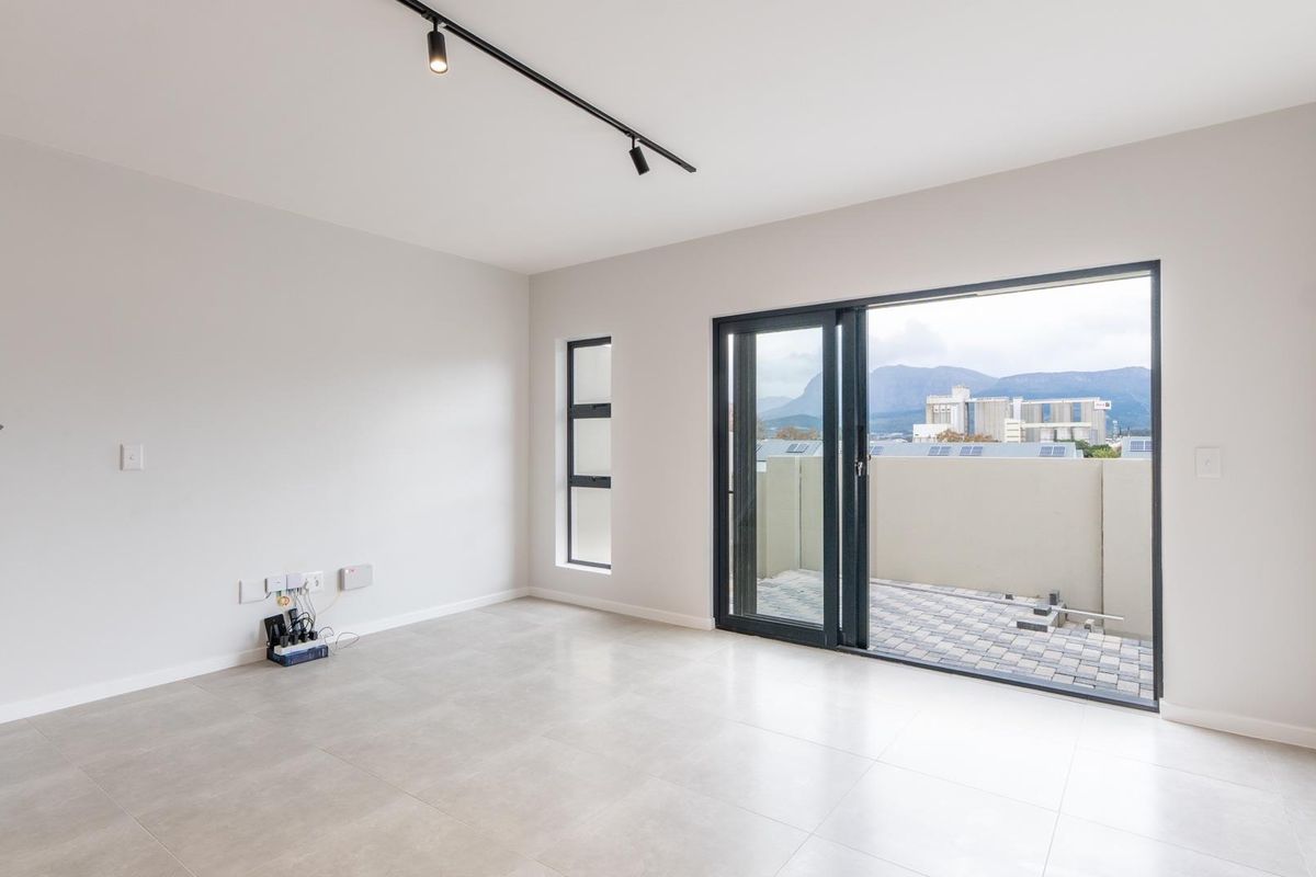 A bright, modern living area featuring neutral floor tiles, track lighting, and large sliding glass doors that open onto a private balcony with mountain views.