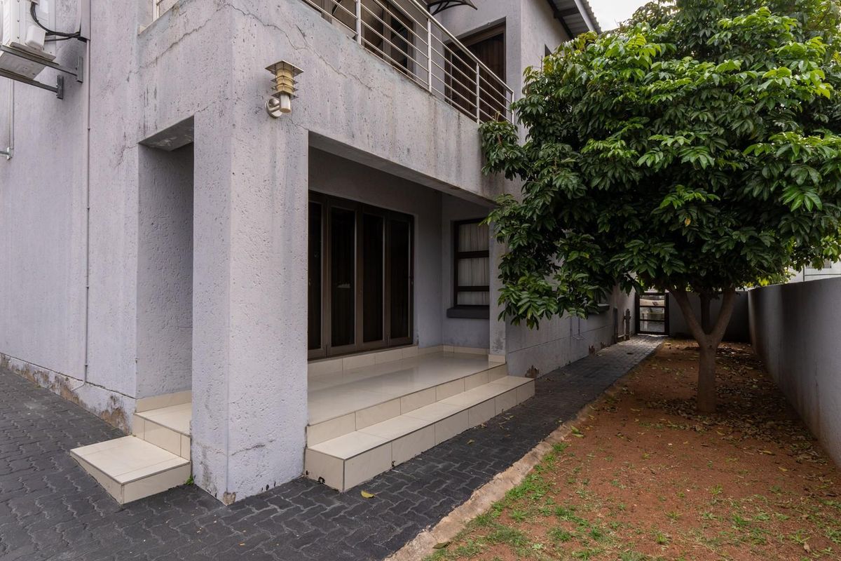 Rear exterior view of the property featuring a covered patio with tiled steps, a balcony above, and a small garden area with a mature tree.