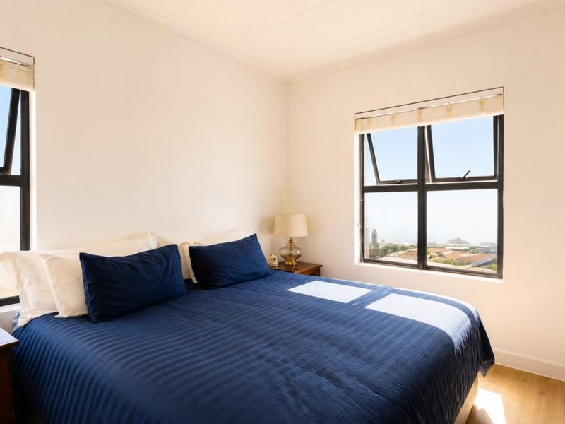 A bright bedroom featuring laminate flooring, built-in wardrobes, and large windows with roller blinds offering natural light and views.