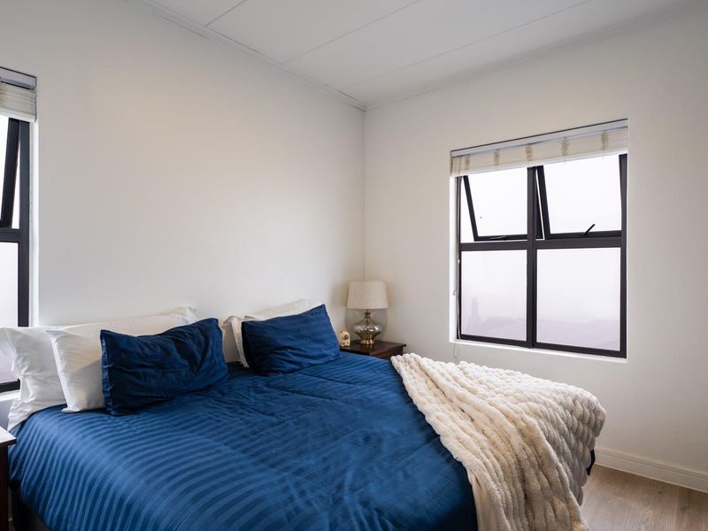 A bright bedroom featuring neutral walls, wood-look flooring, built-in wardrobe storage, and dual windows with fitted roller blinds.