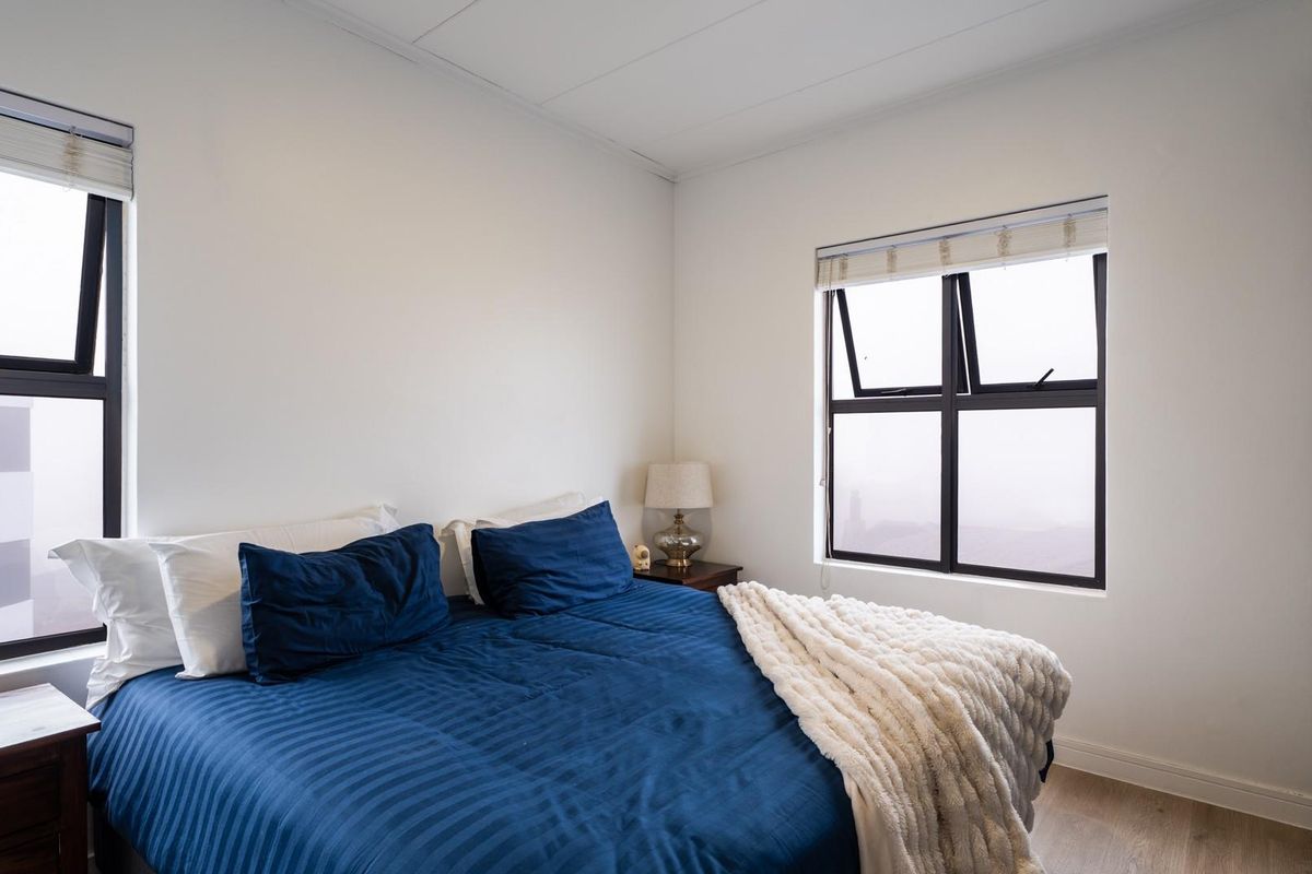 A bright bedroom featuring neutral walls, wood-look flooring, built-in wardrobe storage, and dual windows with fitted roller blinds.