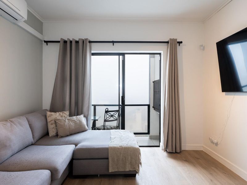 A well-maintained living room featuring laminate flooring, a wall-mounted television, and a sliding glass door providing access to a balcony.