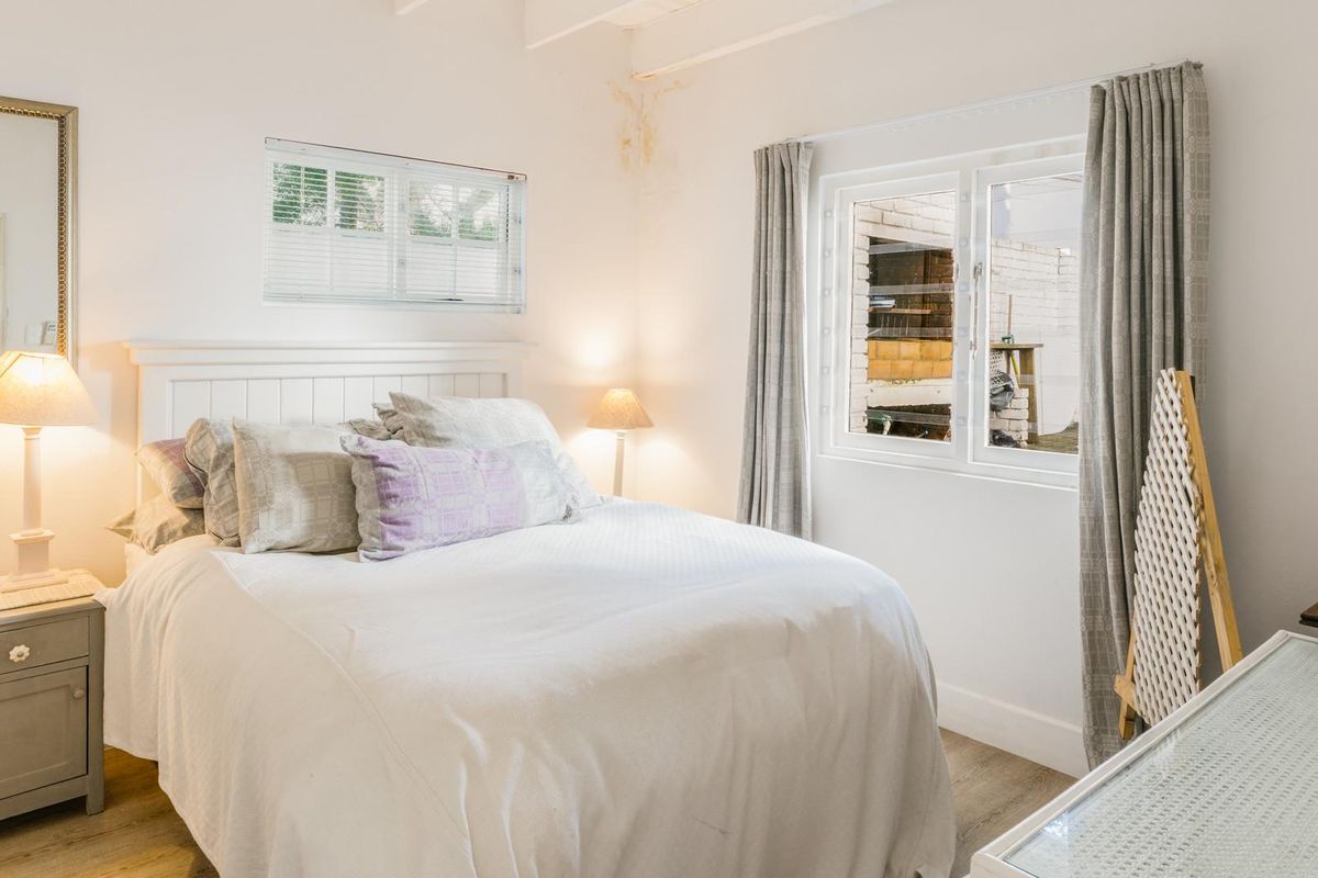 A cozy bedroom featuring laminate flooring, exposed ceiling beams, and dual windows. Note visible water staining on the upper wall requiring attention.