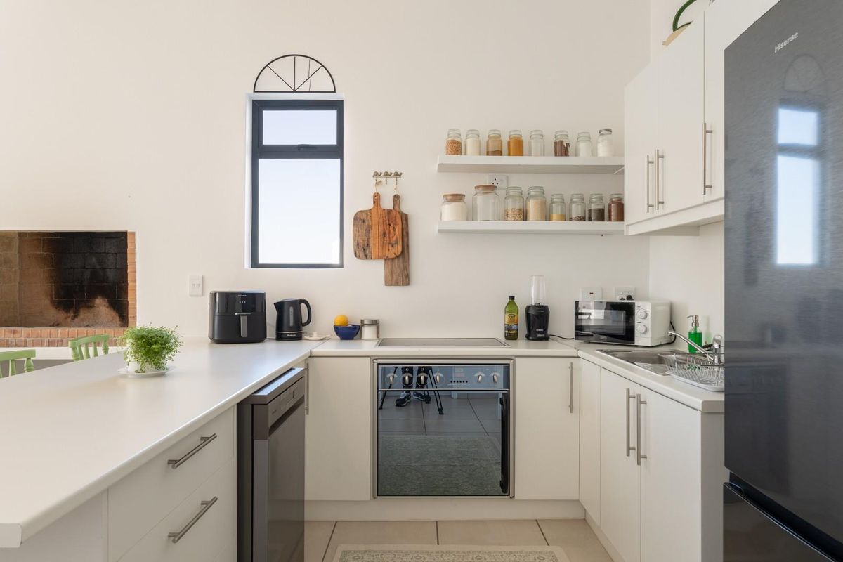 A bright, modern kitchen featuring white cabinetry, integrated oven and hob, floating shelves, and a built-in fireplace in the adjacent dining area.