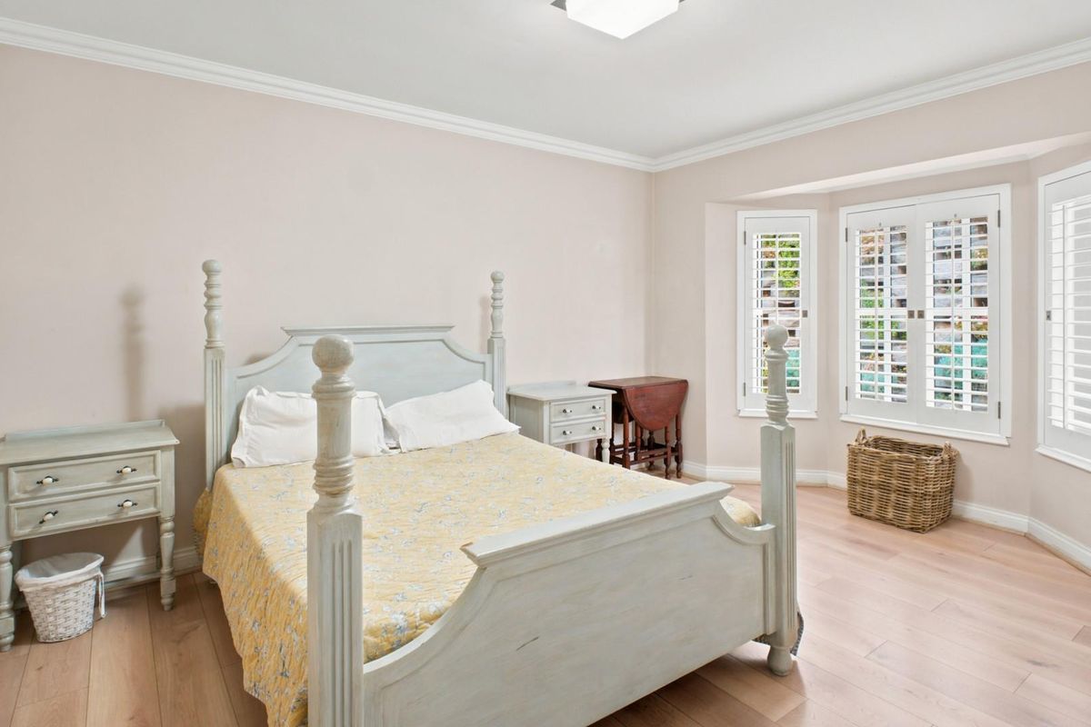 A bright and spacious bedroom featuring light-toned timber flooring, crown molding, and plantation shutters on the bay windows.