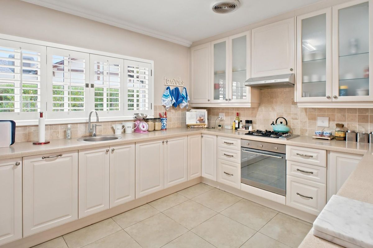 A well-maintained kitchen featuring cream cabinetry, stone-look countertops, tiled backsplash, built-in oven, gas hob, extractor fan, and plantation shutters.