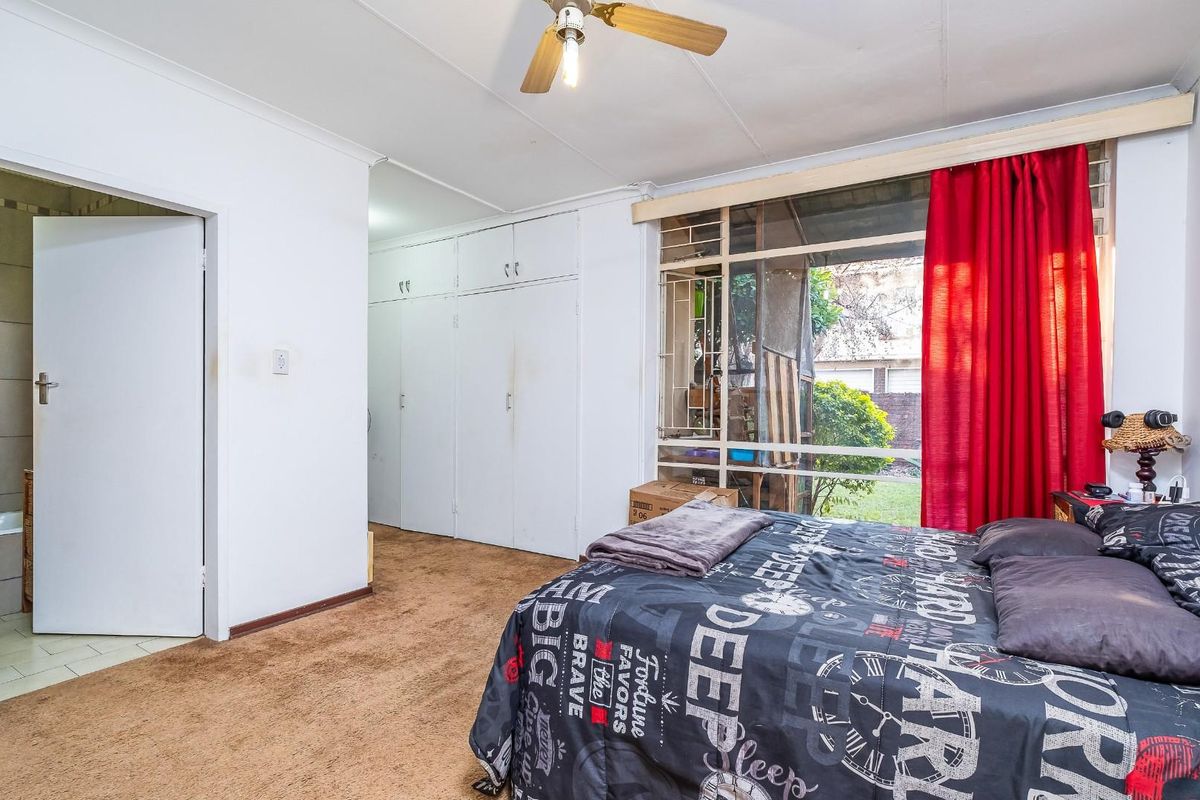 A bedroom featuring built-in cupboards, a ceiling fan, and large windows with security bars, providing direct access to an en-suite bathroom.