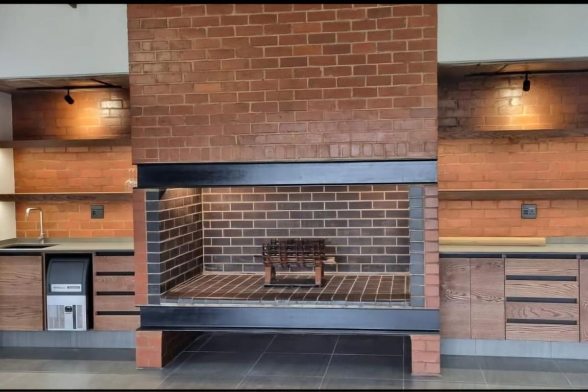 A modern indoor braai area featuring a large brick fireplace, built-in wooden cabinetry, floating shelves, and a stainless steel under-counter ice maker.