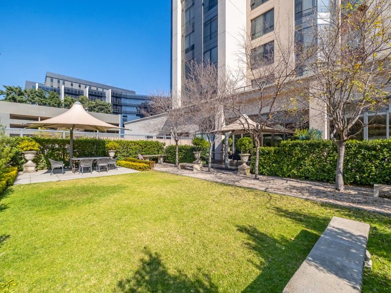 A well-maintained communal garden area featuring a manicured lawn, paved patio sections, and established hedge landscaping within an apartment complex setting.