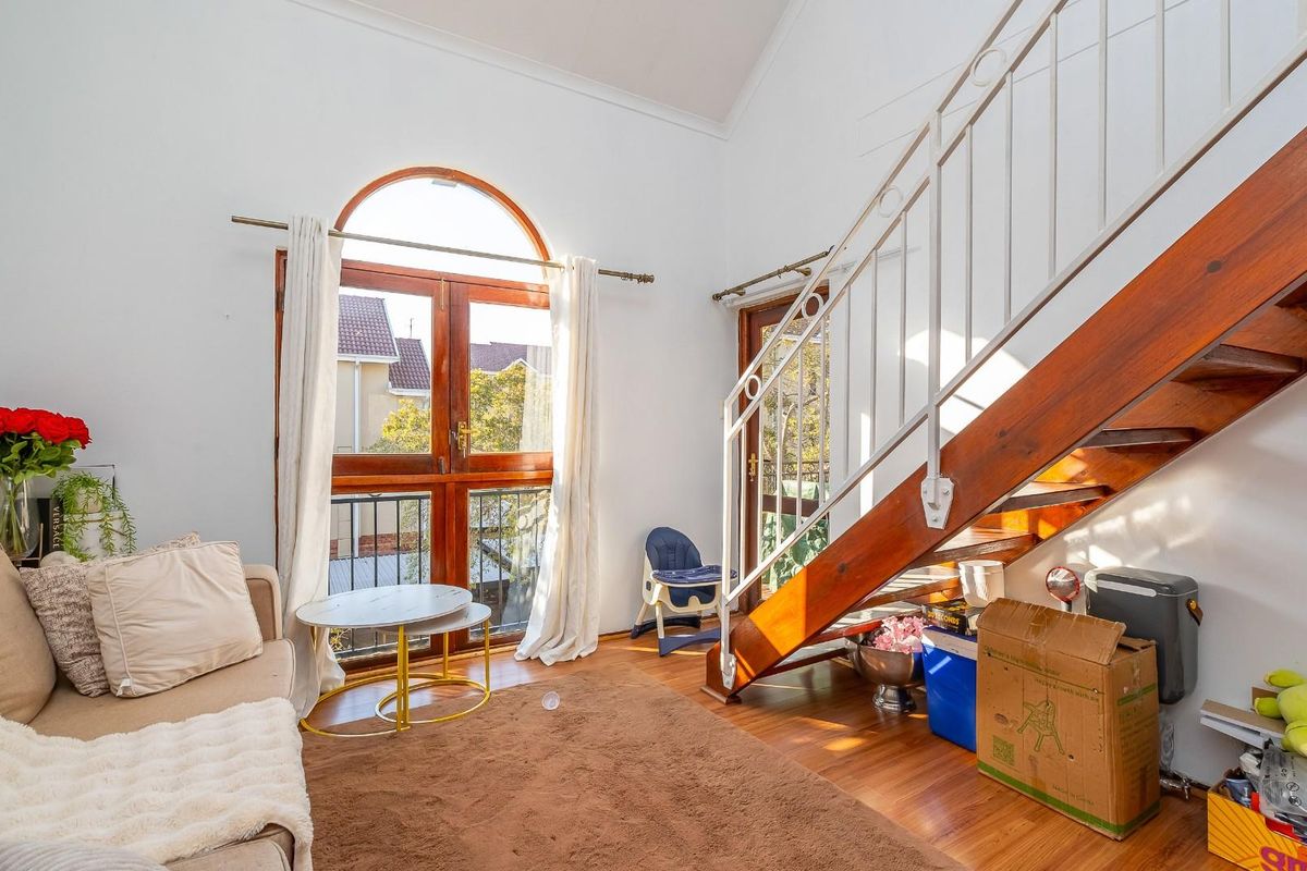 A bright living area featuring timber flooring, a prominent timber staircase with metal balustrade, and dual access to an outdoor balcony through arched timber doors.