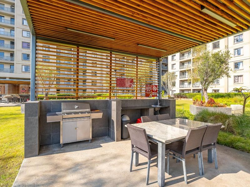 Outdoor communal entertainment area featuring a built-in braai station, stone-clad counter, sink, and timber-slatted pergola roof structure set within landscaped apartment grounds.