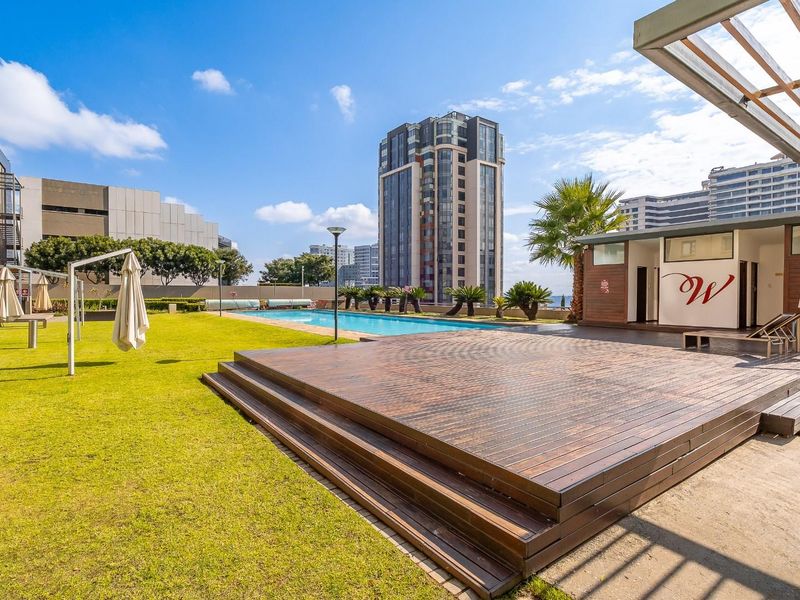 A well-maintained communal pool area featuring a timber deck, manicured lawn, and modern building architecture in an urban setting.