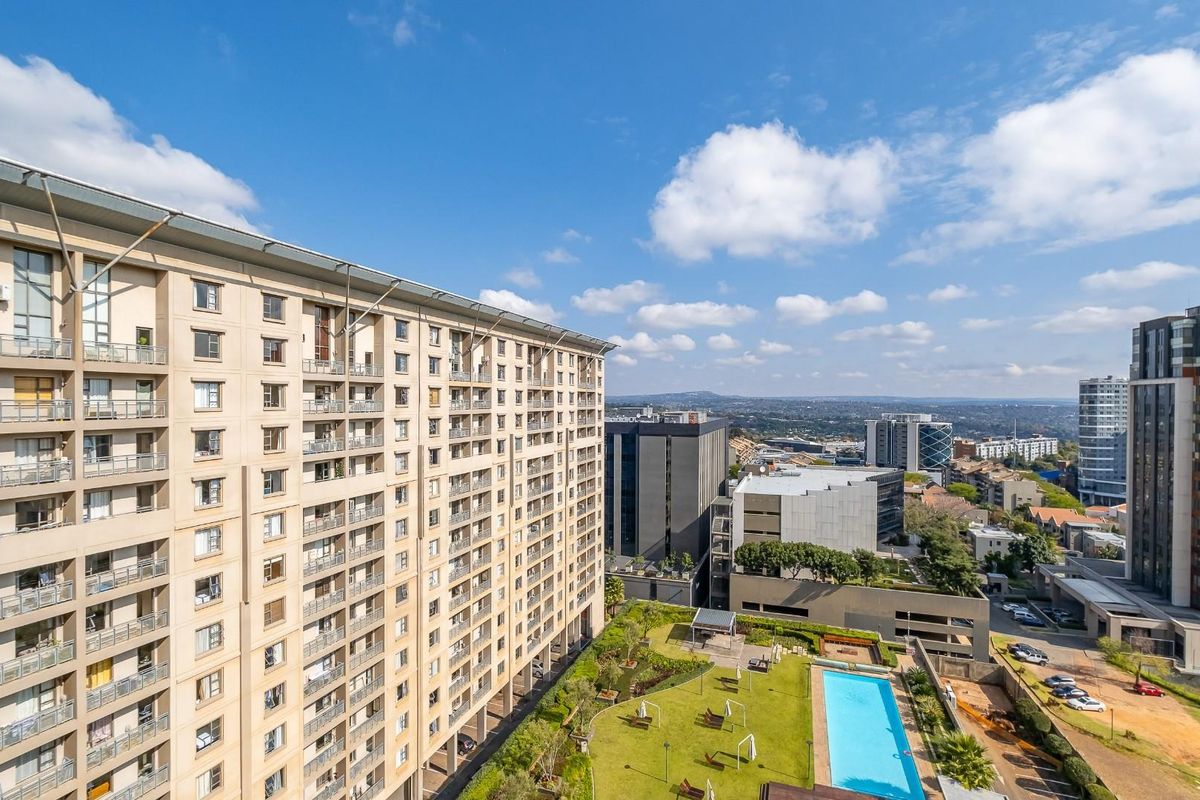 Elevated aerial view of a modern apartment complex featuring a landscaped garden, swimming pool, and surrounding urban cityscape.