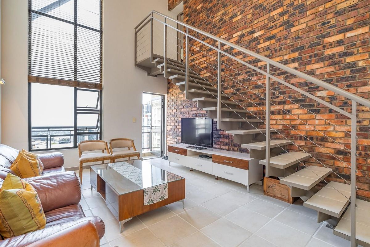 Spacious double-volume living area featuring an exposed brick feature wall, large windows for natural light, and a modern floating staircase.