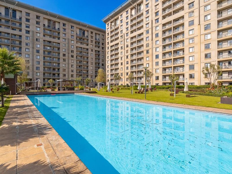 A well-maintained communal swimming pool area set within a landscaped garden, surrounded by modern apartment building facades.