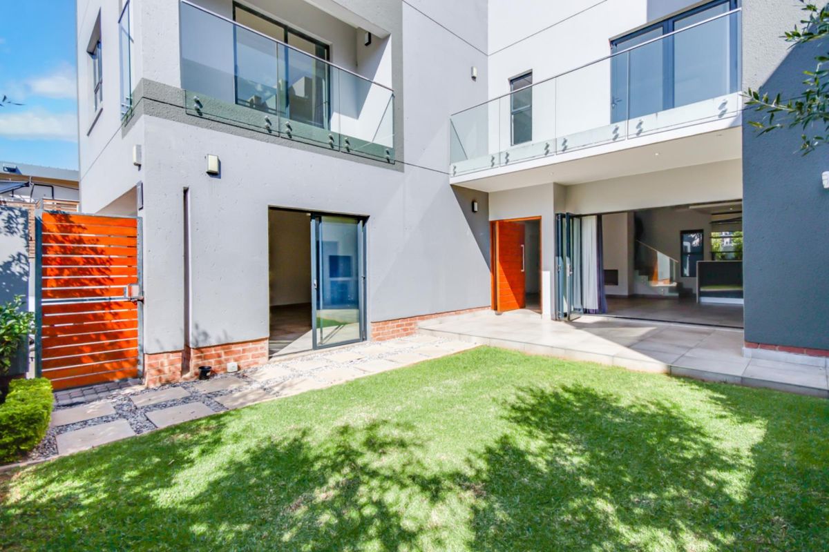 A modern townhouse rear exterior featuring a manicured lawn, tiled patio area, glass balustrades on upper balconies, and seamless indoor-outdoor flow through sliding and folding doors.