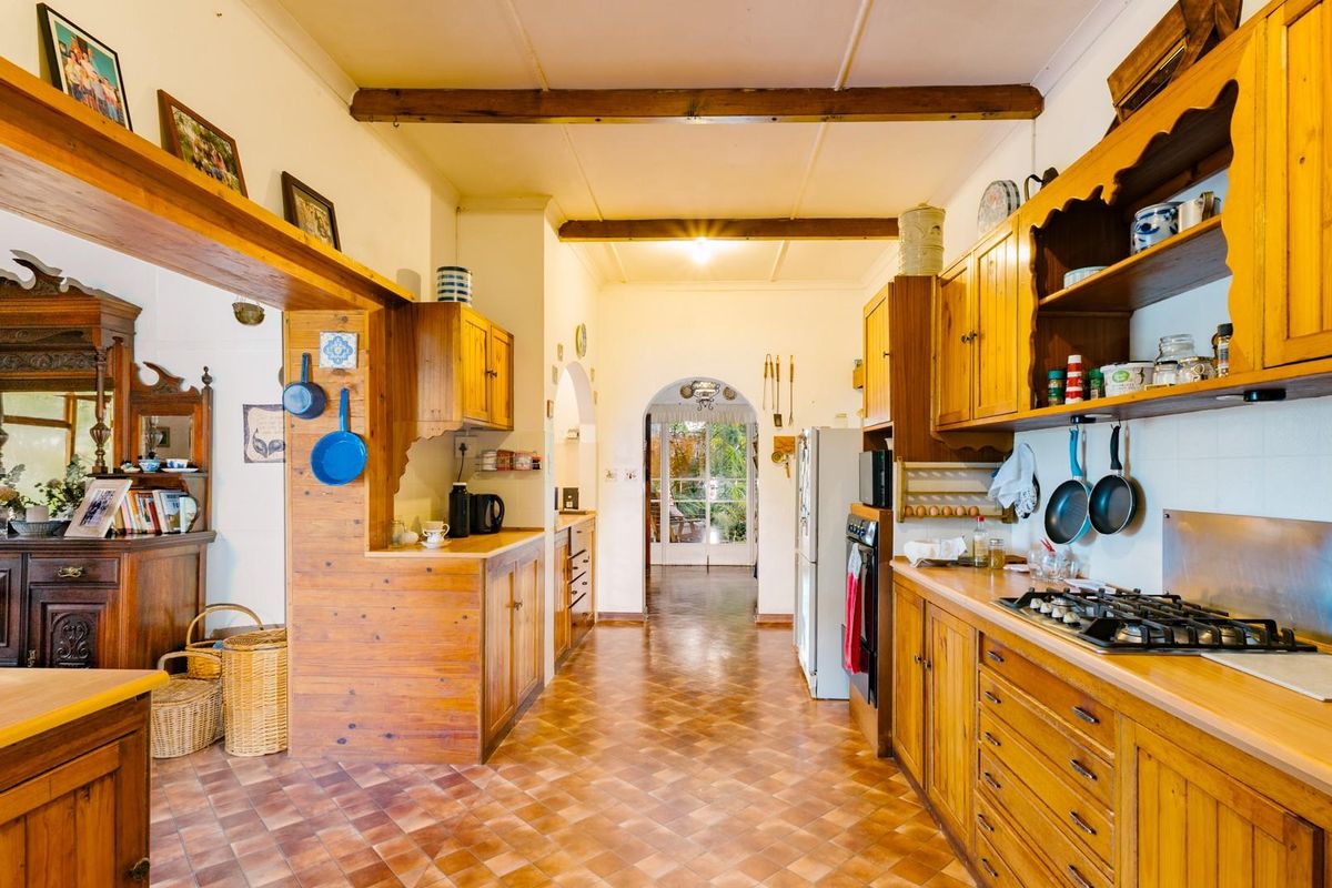 A rustic farmhouse-style kitchen featuring extensive timber cabinetry, exposed ceiling beams, and a gas cooktop, leading through an arched doorway to the garden.
