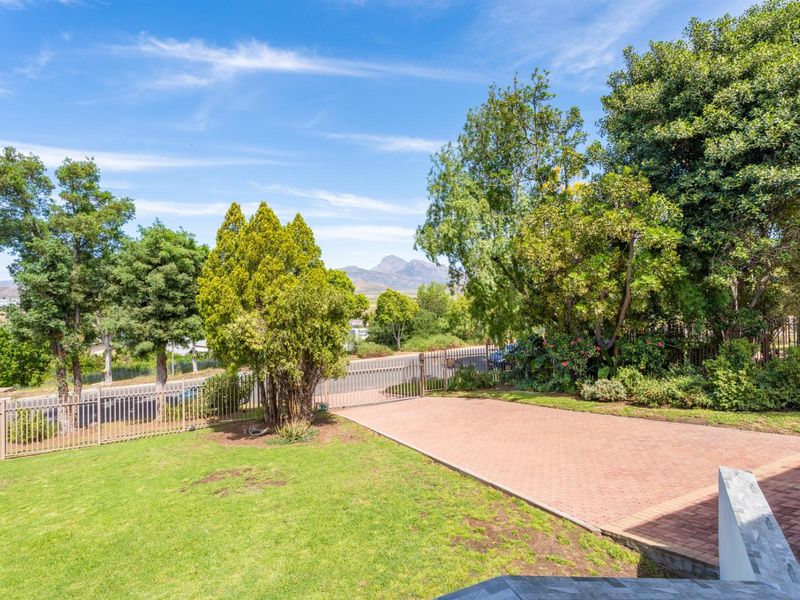 A well-maintained front exterior featuring a paved driveway, metal perimeter fencing, and a landscaped garden with mountain views in the background.