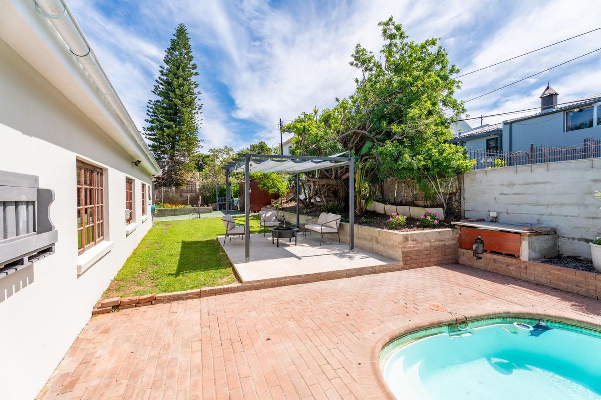 Rear garden area featuring a swimming pool, brick paved patio, pergola, and built-in braai, set against a landscaped retaining wall.