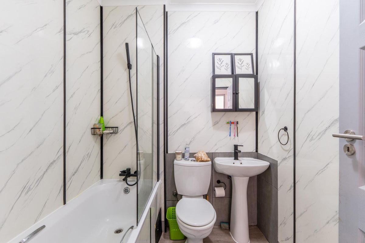 A compact bathroom featuring a bathtub with a glass shower screen, a pedestal sink, and a toilet, finished with marble-effect wall paneling.