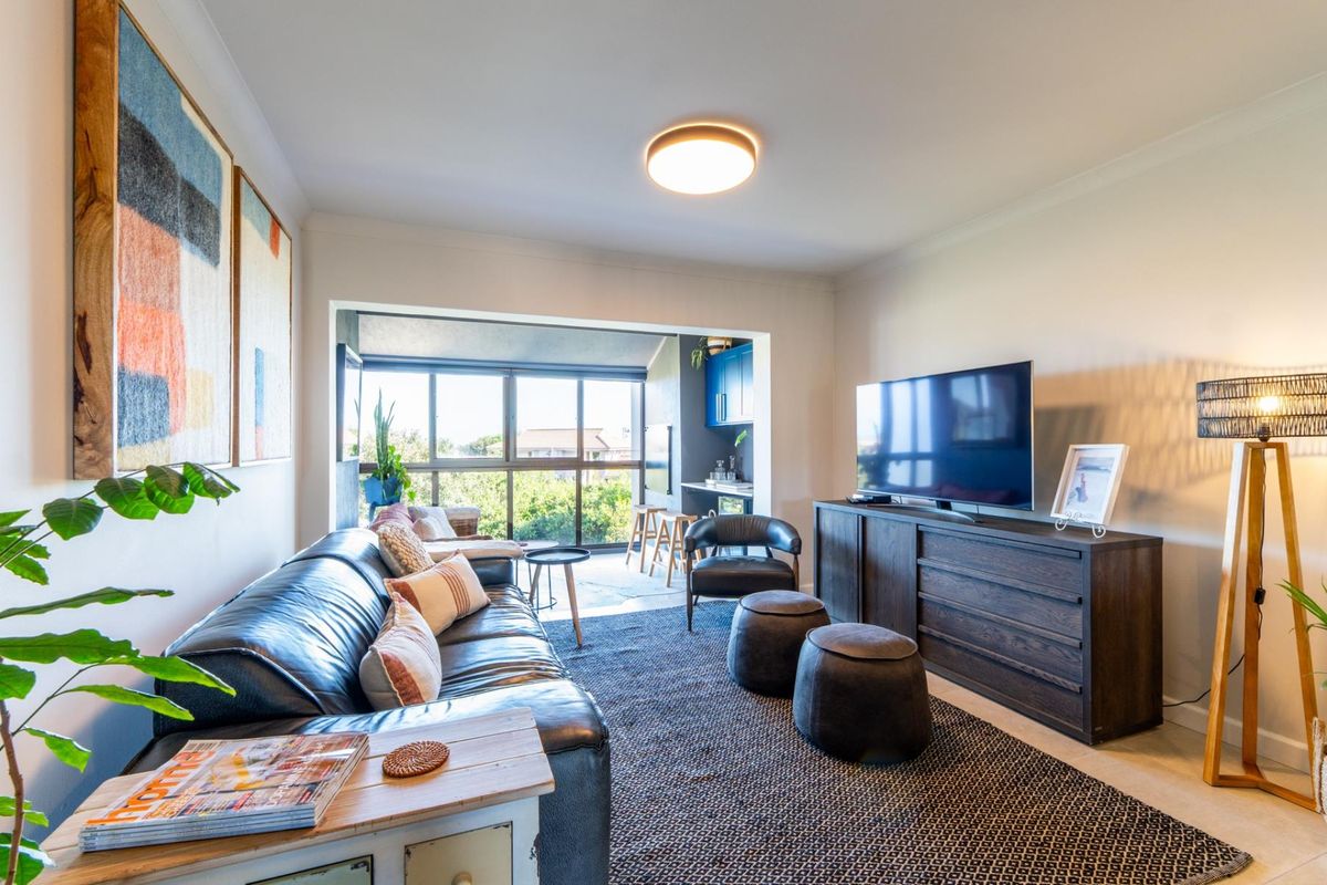A comfortable living room featuring neutral walls, recessed lighting, and a large window area that connects to a secondary kitchen or bar space.