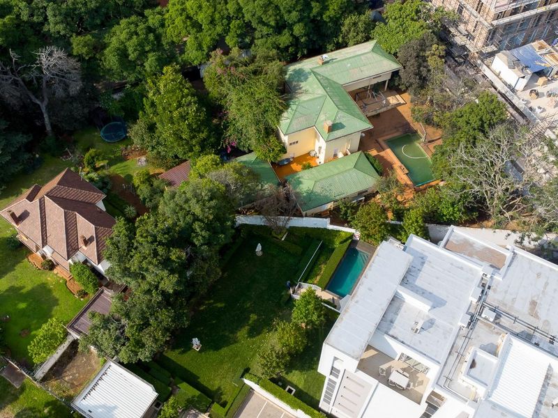 An aerial view of a residential property featuring a large garden, swimming pool, and multiple roof structures surrounded by mature trees.