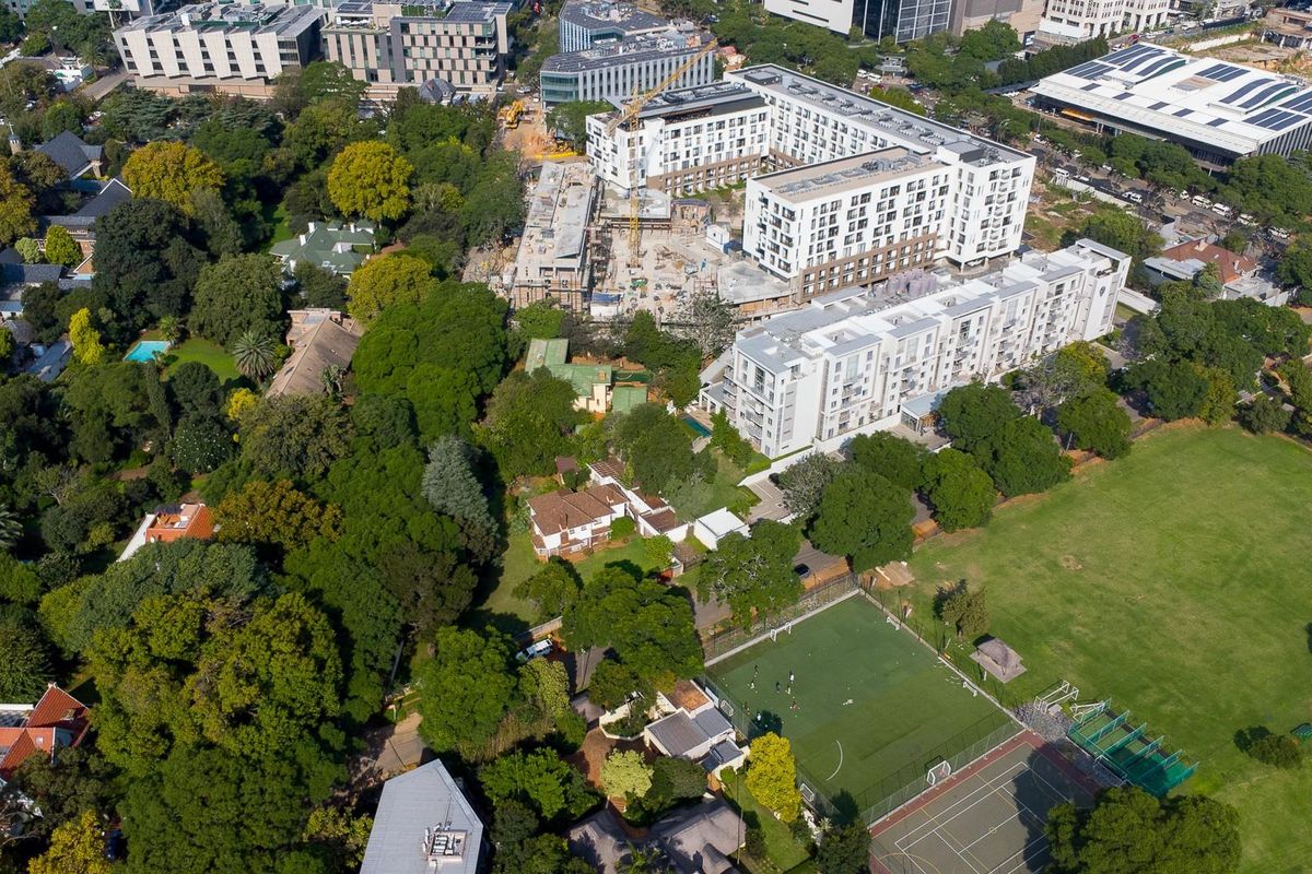 An aerial view of a residential area featuring mature trees, nearby apartment complexes, and recreational sports facilities including a field and tennis court.