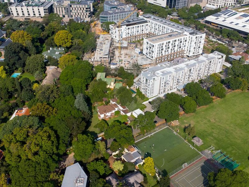 An aerial view of a residential area featuring mature trees, nearby apartment complexes, and recreational sports facilities including a field and tennis court.