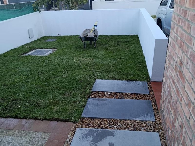 A compact, low-maintenance garden area featuring a small lawn, stone paver pathway, and clean boundary walls.