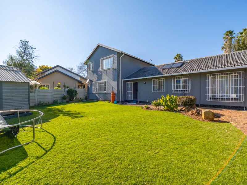 Rear view of the property featuring a spacious lawn, paved walkway, and a grey-painted exterior with security bars on windows.