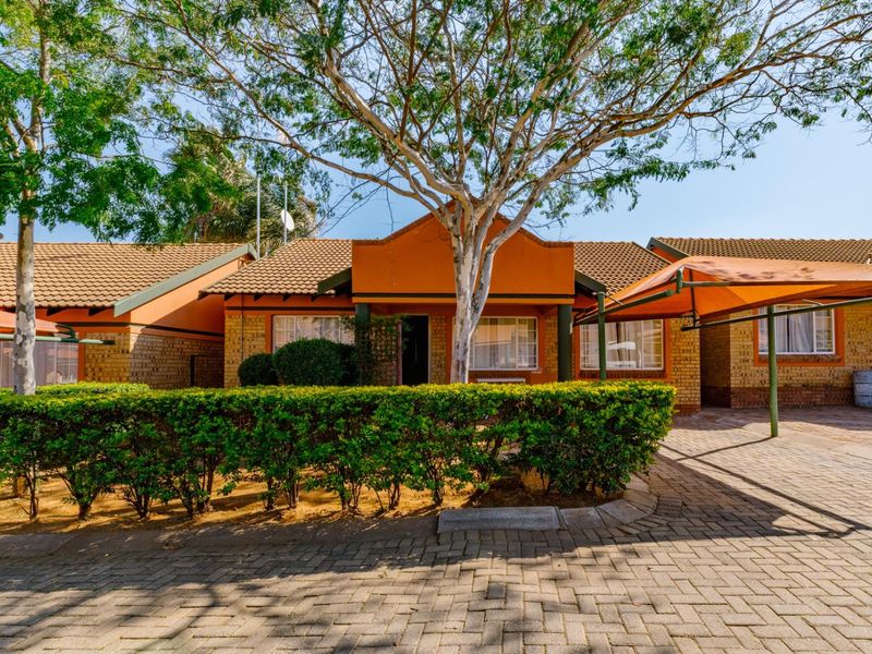 Front view of a townhouse featuring a brick facade, tiled roof, paved driveway with a carport, and a well-maintained hedged garden.